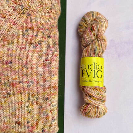 Multicolored 'firefly' yarn skein with 'studio EVIG' label next to a knitted garment on a green and white background