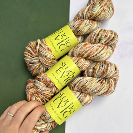 Three skeins of multicoloured yarn with 'Studio EVIG' labels held by a hand against a green and white background.